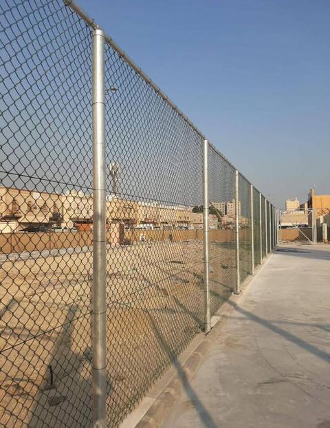 سور شبك مجلفن مرتفع حول ملعب رياضي بجانب رصيف داخل حي سكني