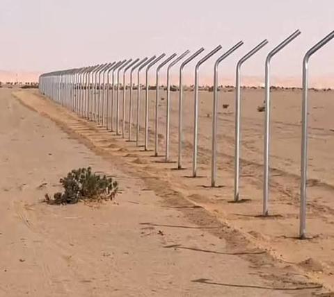 صف طويل من أعمدة سياج مجلفنة مائلة الرأس ممتد في أرض صحراوية لتسوير محيط مزرعة